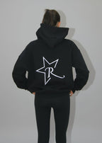 Person wearing a black hoodie with a logo on the back, standing against a white backdrop.