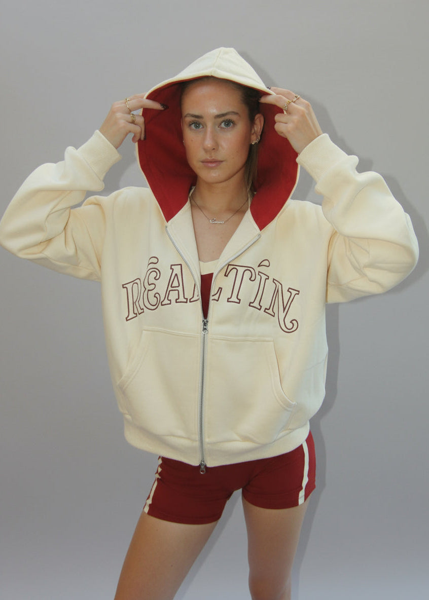 Person wearing a cream hoodie with red lining and 'REALTIN' text, standing against a plain background.